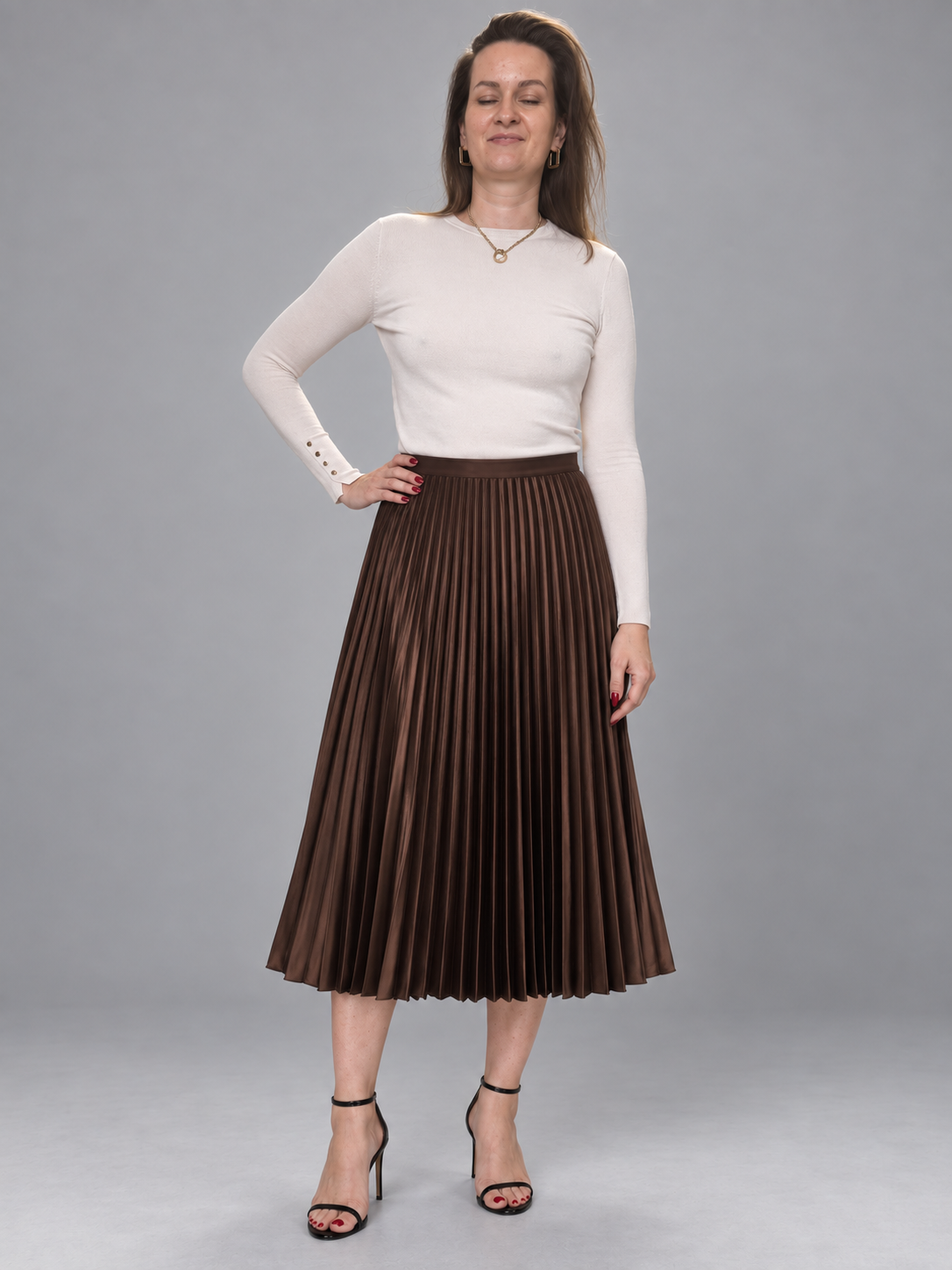 A woman modeling a brown pleated midi skirt with a white long-sleeve top and black high-heeled sandals, standing against a solid gray studio background.
