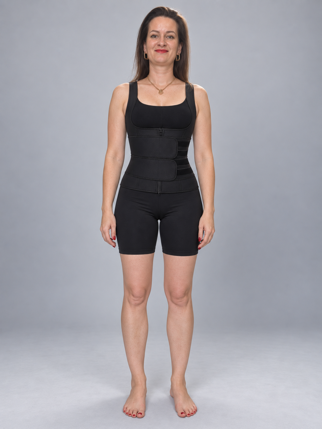A woman stands barefoot against a light gray studio background wearing a black waist trainer vest with matching black fitted shorts.