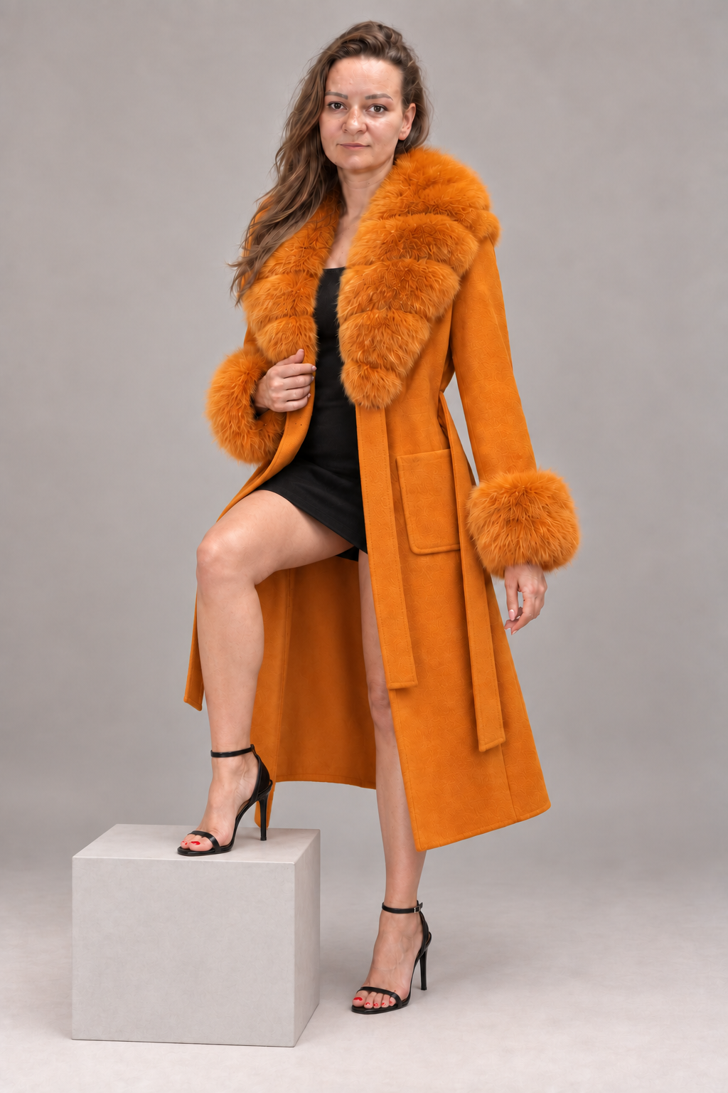 A woman models a long orange suede-like coat with a voluminous orange fur collar and matching fur cuffs, worn over a black dress, standing on a light gray pedestal against a neutral gray studio background.