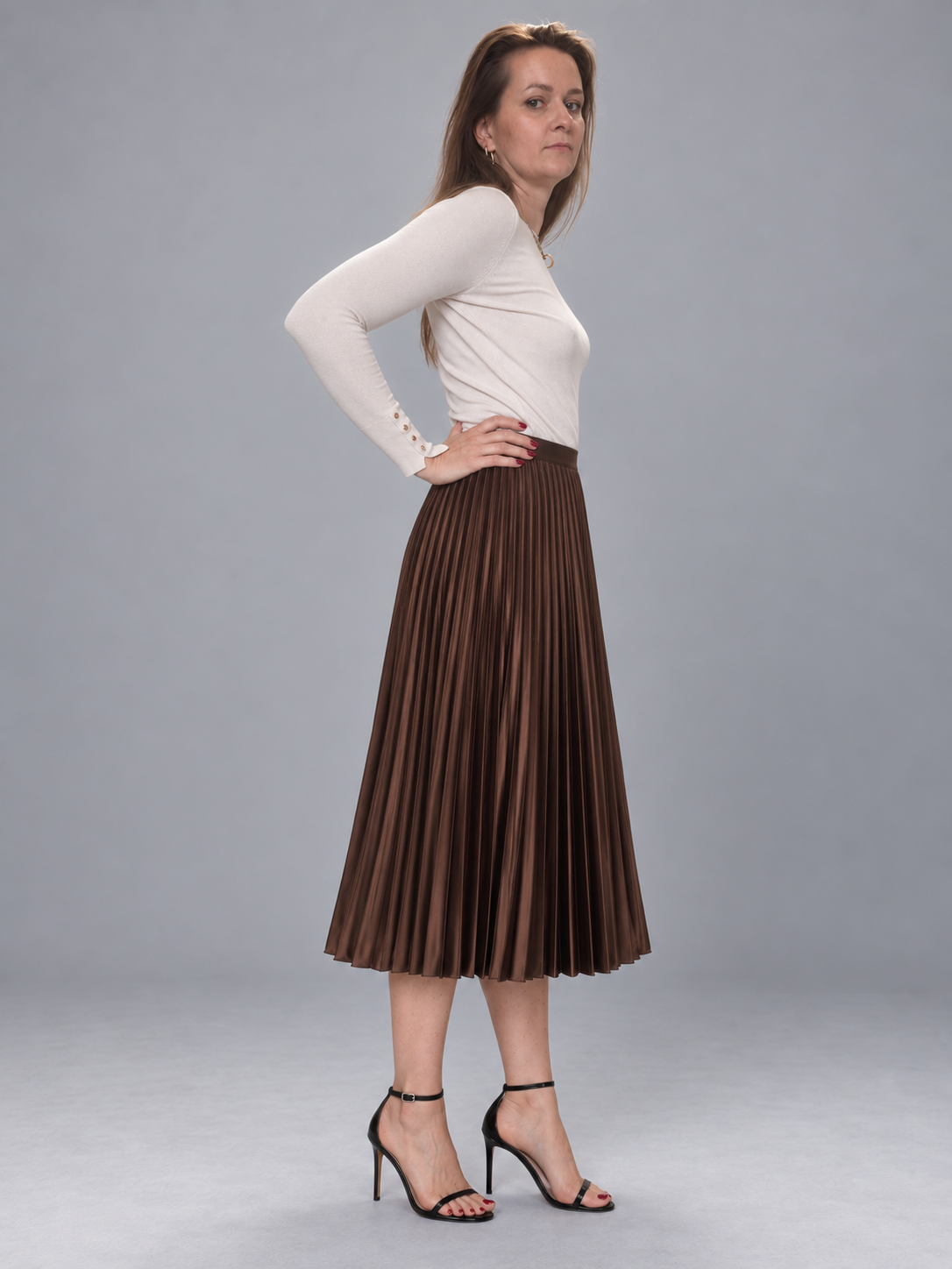 A woman modeling a long-sleeve white top and a brown pleated midi skirt, paired with black high-heeled sandals, against a solid gray studio background.