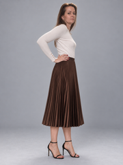 A woman modeling a long-sleeve white top and a brown pleated midi skirt, paired with black high-heeled sandals, against a solid gray studio background.