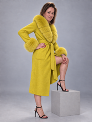 A woman models a bright yellow wool coat with a fluffy yellow fur collar and cuffs, paired with black high-heeled sandals, posing on a gray block against a gray studio background.