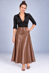 A woman modeling a brown faux-leather A-line midi skirt paired with a black button-down shirt, standing against a light gray studio background.