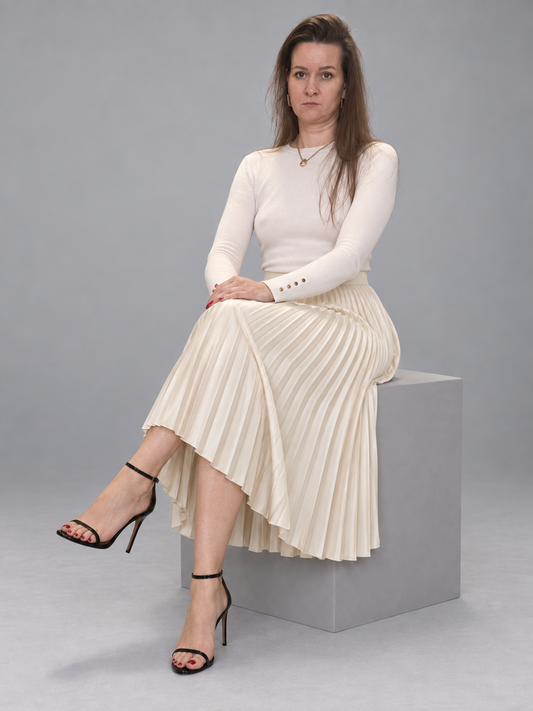 A woman sits on a gray pedestal wearing a cream long-sleeve knit top and matching pleated midi skirt, paired with black strappy high-heel sandals, against a solid gray studio background.