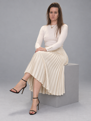 A woman sits on a gray pedestal wearing a cream long-sleeve knit top and matching pleated midi skirt, paired with black strappy high-heel sandals, against a solid gray studio background.