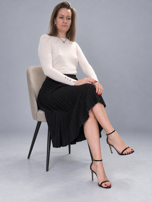 A woman sits on a light gray upholstered chair against a plain gray studio background, wearing a fitted white long-sleeve top, a black pleated midi skirt, and black strappy high-heel sandals.