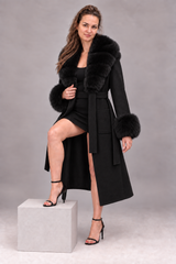 A woman models a long black coat with a luxurious black fur collar and matching fur-trimmed cuffs, paired with a black dress and black high-heeled sandals, standing on a light gray pedestal against a neutral gray studio background.
