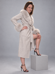 A woman models a light beige faux-suede wrap coat with fluffy light gray fur trim at the collar, cuffs, and waist, paired with black high-heeled sandals, standing on a gray pedestal against a gray studio background.