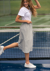 Woman on a tennis court wearing a white t-shirt and gray skirt.