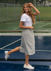 Woman standing on a tennis court wearing a white t-shirt and light gray skirt.