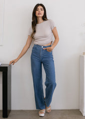 Woman wearing a beige top and blue jeans standing against a white wall.