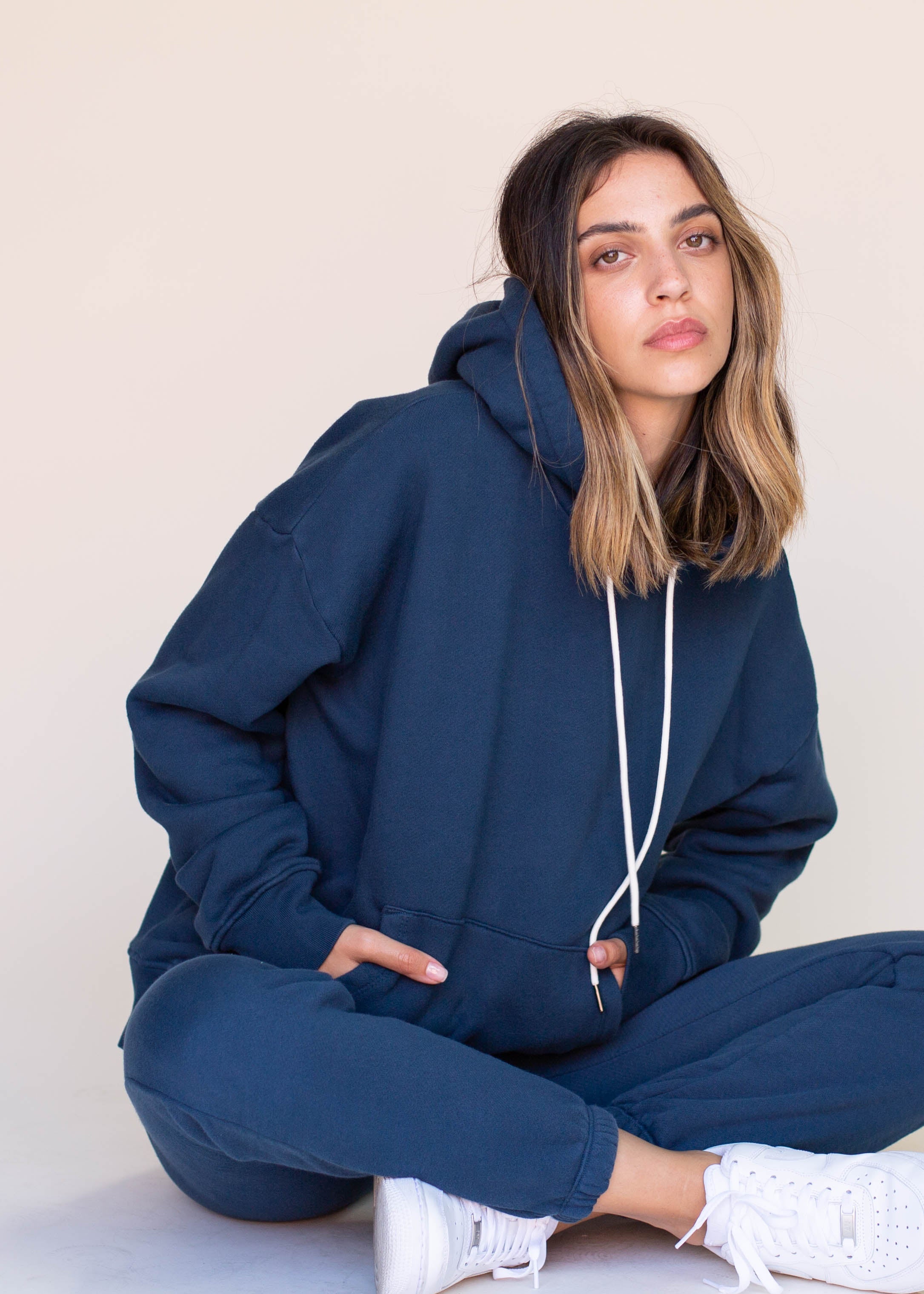 Person wearing a navy blue hoodie and pants sitting on the floor against a plain background