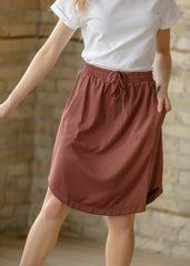 Person wearing a white shirt and brown skirt against a stone wall background