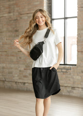 Woman wearing a white t-shirt and black skirt with a black bag, standing in a room with brick walls and large windows.