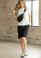 Woman wearing a white t-shirt, black shorts, and white sneakers, holding a black waist bag in a room with brick walls.