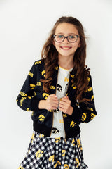 Young girl wearing a black jacket with yellow patterns, holding a white cup.