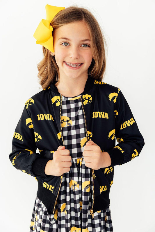 Young girl wearing a black jacket with 'Iowa' pattern and checkered dress, smiling.