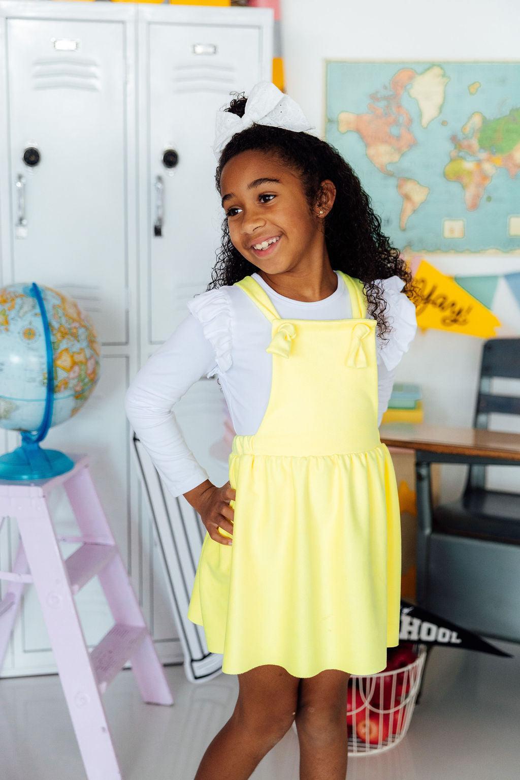 Young girl in a yellow dress standing in a classroom setting with educational items around.