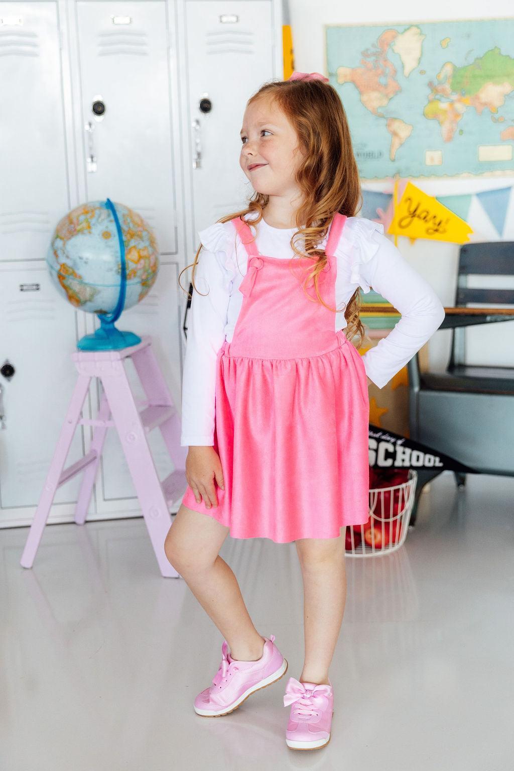 Young girl in a pink dress standing in a classroom setting with educational items around.