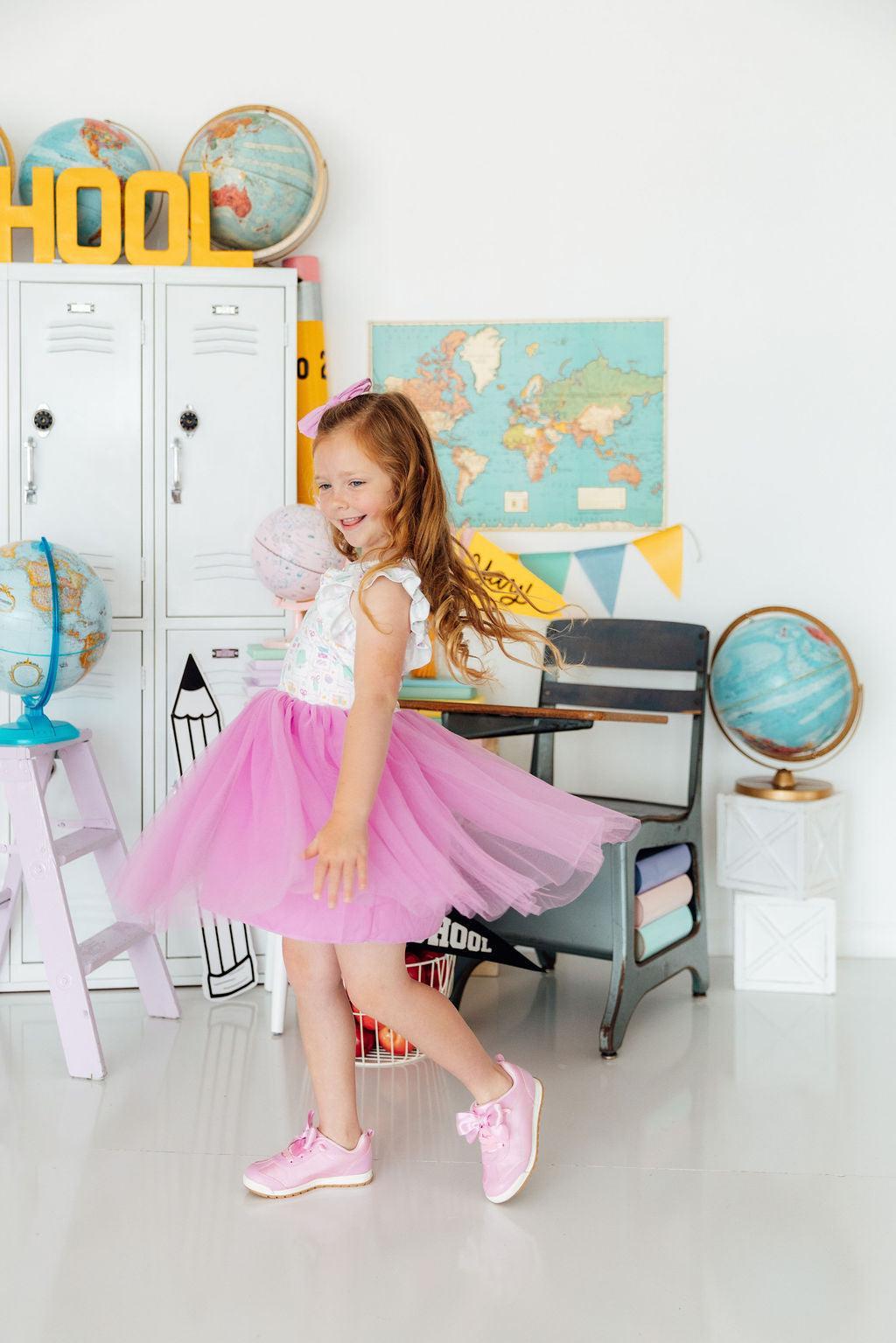 Young girl in a pink tutu standing in a classroom setting with educational decor.
