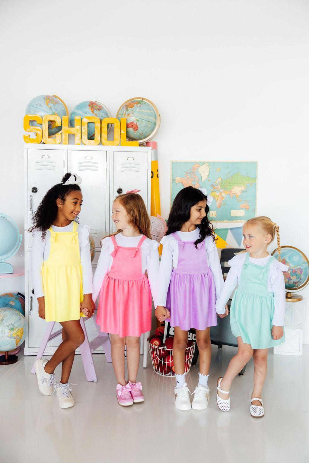 Four children in colorful dresses standing in a classroom setting with educational posters and a 'School' sign.