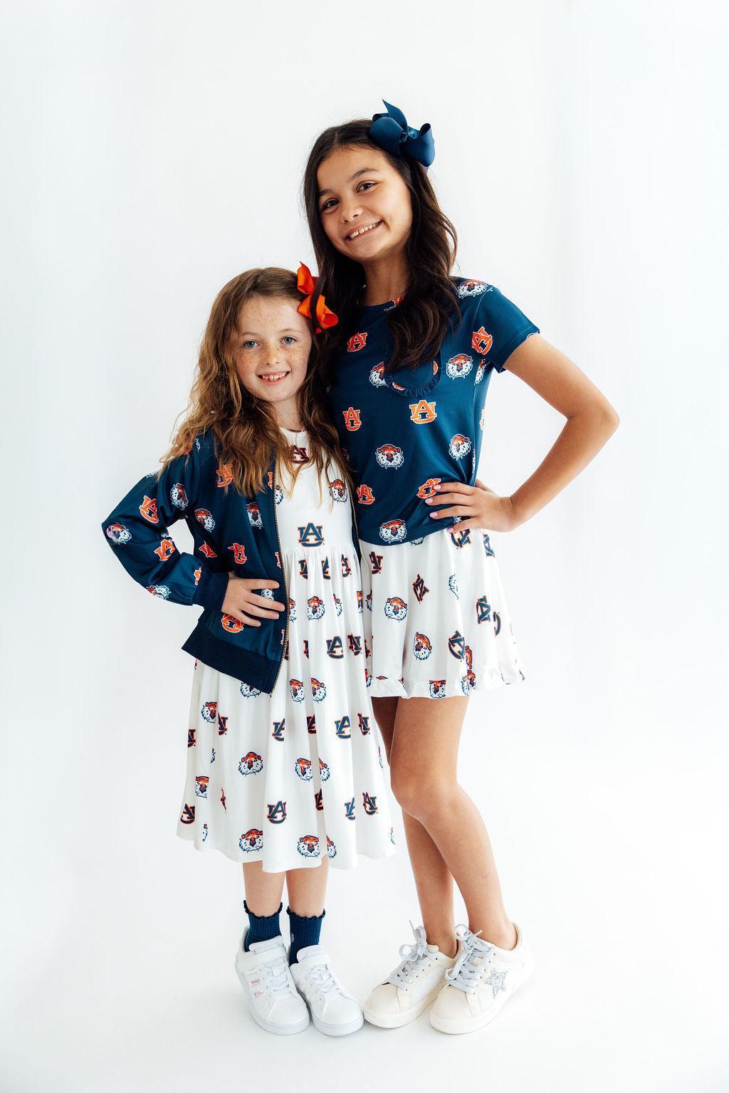 Two young girls modeling matching navy blue and white patterned outfits against a plain white studio background. The girl on the right wears a navy short-sleeve top with a white skirt featuring a small repeating character print, and the girl on the left wears a matching navy cardigan over a white dress.