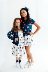 Two young girls modeling matching navy blue and white patterned outfits against a plain white studio background. The girl on the right wears a navy short-sleeve top with a white skirt featuring a small repeating character print, and the girl on the left wears a matching navy cardigan over a white dress.