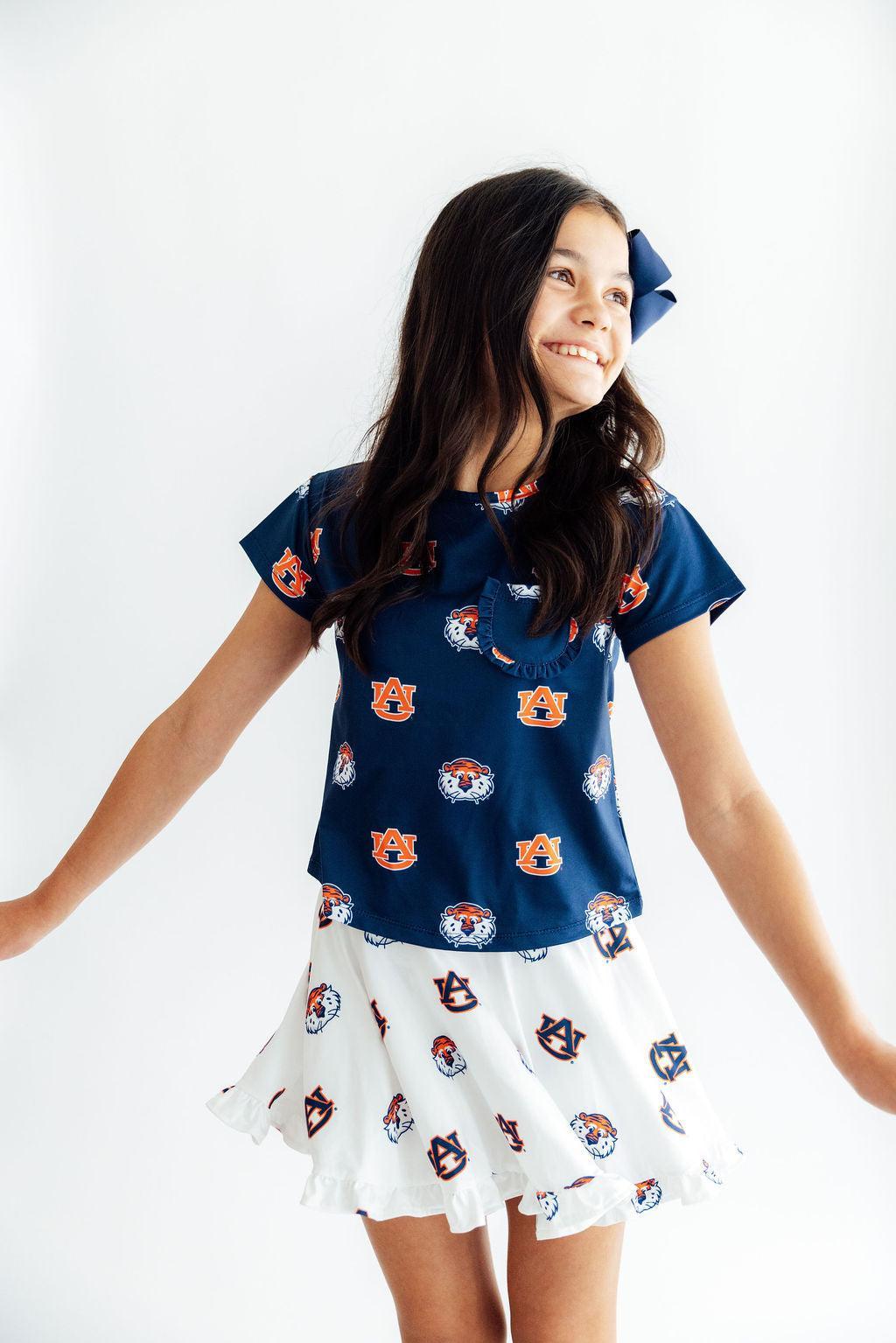 A young girl smiling and looking to the side, wearing a navy blue short-sleeve top with an orange and white tiger face pattern, paired with a white ruffled skirt with matching animal prints, and a navy hair bow.