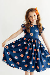 A young girl with long light brown hair wearing a navy blue dress with an orange and white tiger face pattern, holding out her skirt to show its volume. She is smiling and posing against a plain white studio background.