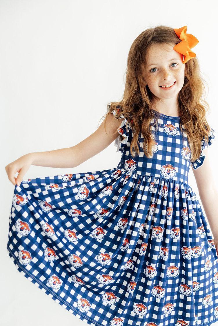 A young girl with long light brown hair wearing a blue and white gingham dress with a repeating pattern of cartoon dog faces, holding out one side of the skirt to show its volume. She is smiling and posing against a plain white background.