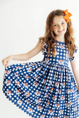 A young girl with long light brown hair wearing a blue and white gingham dress with a repeating pattern of cartoon dog faces, holding out one side of the skirt to show its volume. She is smiling and posing against a plain white background.
