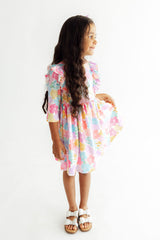 A young girl with long dark hair stands in profile wearing a pastel multicolor floral tiered dress with ruffled sleeves and white sandals, photographed against a plain white studio background.