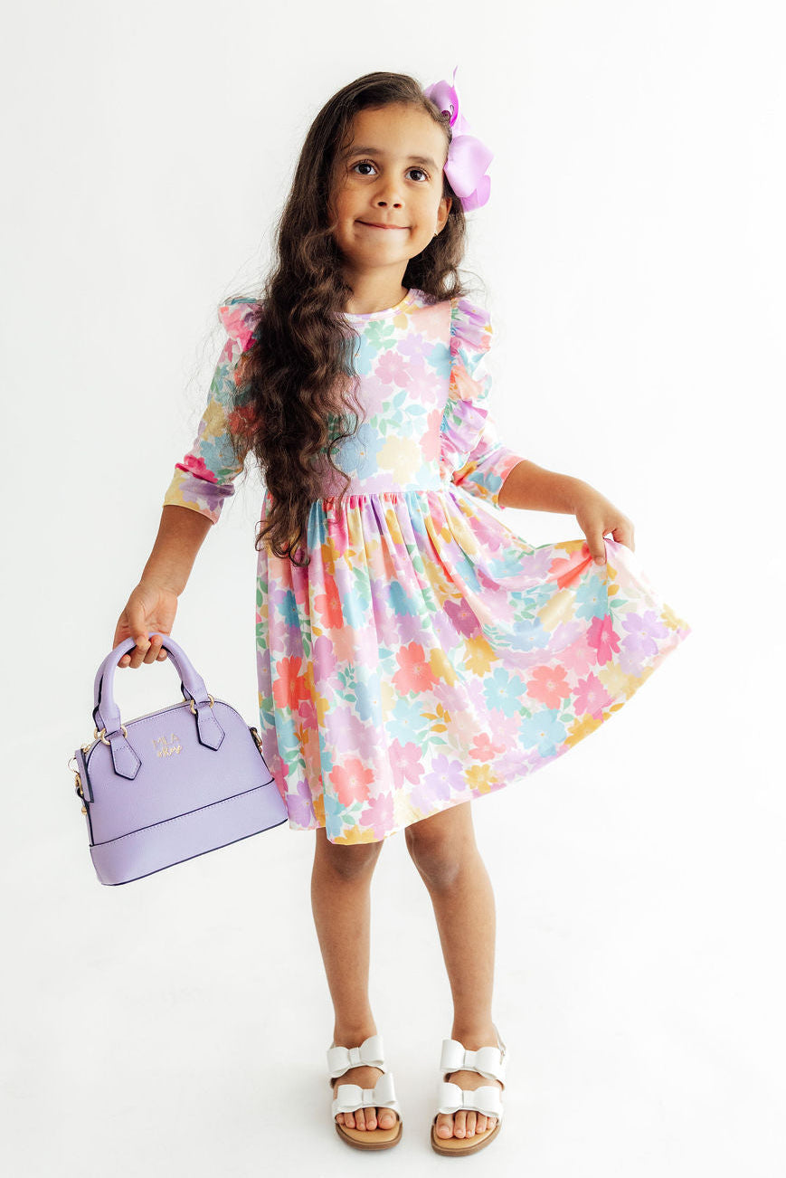 A young girl with long dark hair wearing a pastel multicolor floral dress, white sandals, and a purple flower hair clip, holding a lavender handbag against a plain white background.