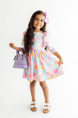 A young girl with long dark hair wearing a pastel multicolor floral dress, white sandals, and a purple flower hair clip, holding a lavender handbag against a plain white background.