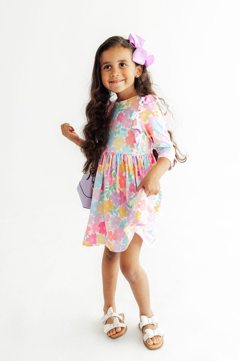 A young girl with long dark hair wearing a pastel multicolor floral print dress, a pink bow, and white sandals, standing against a plain white studio background.