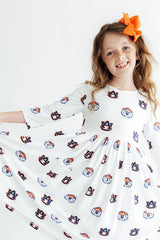 A young girl smiling and twirling in a white long-sleeve dress with an all-over Auburn Tigers logo print, holding out her skirt to show the pattern. She has a bright orange hair bow and is photographed against a clean white background.