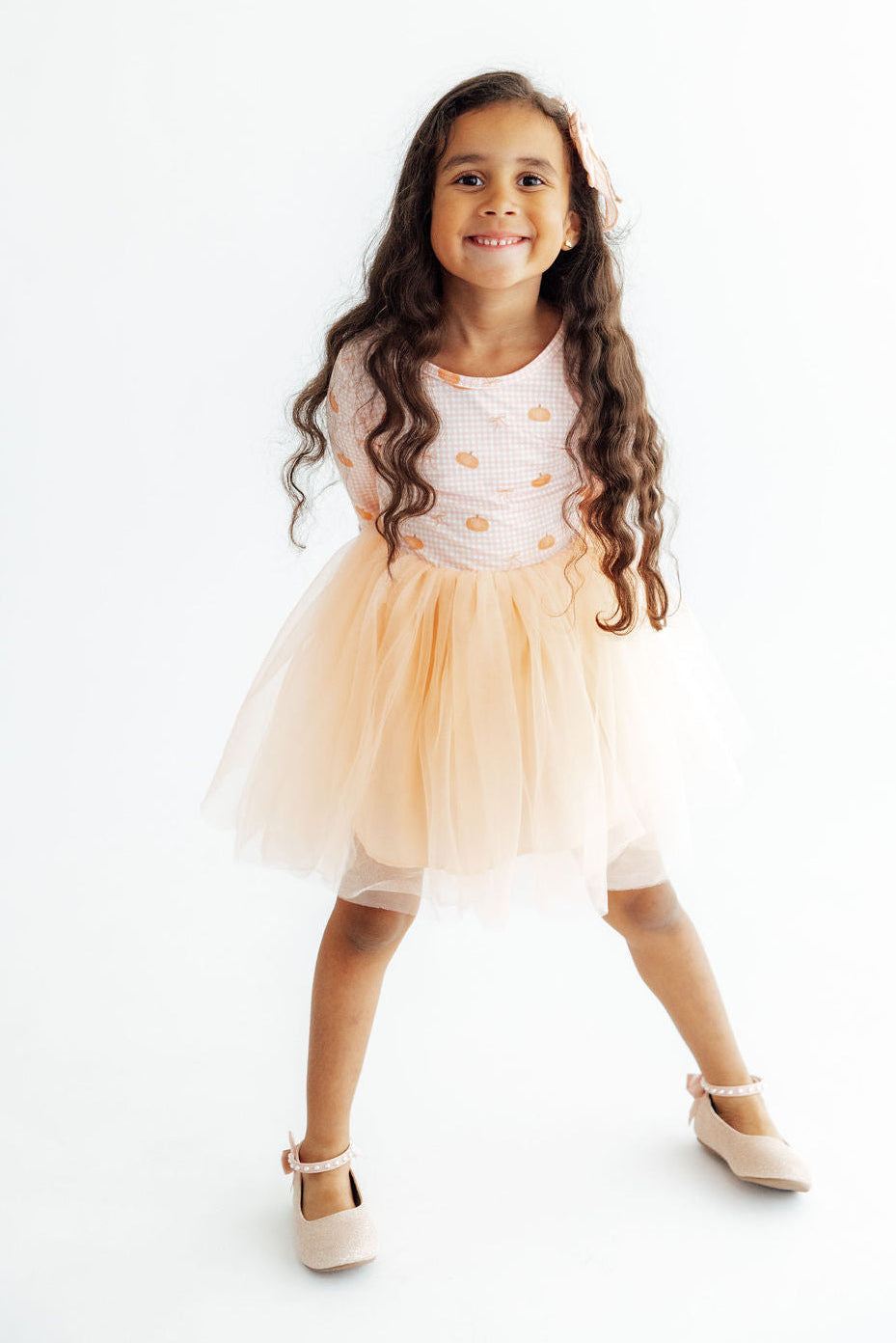 A young girl with long, wavy brown hair stands in a playful pose wearing a light peach tulle tutu dress with a white polka-dot bodice and beige Mary Jane shoes, set against a plain white studio background.