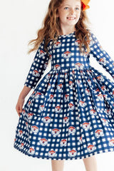A young girl with long wavy light brown hair wearing a blue and white gingham dress with a repeating pattern of cartoon dog faces, posed against a plain white background.