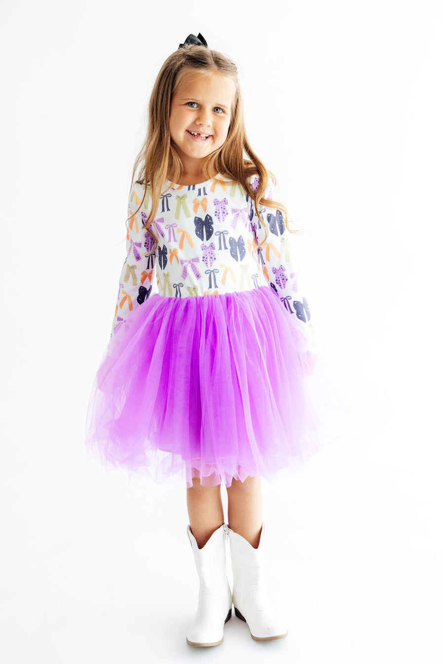 A young girl wearing a long-sleeve white dress with a colorful cactus print and a bright purple tulle tutu skirt, paired with white cowboy boots, standing against a plain white background.
