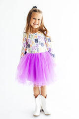 A young girl wearing a long-sleeve white dress with a colorful cactus print and a bright purple tulle tutu skirt, paired with white cowboy boots, standing against a plain white background.