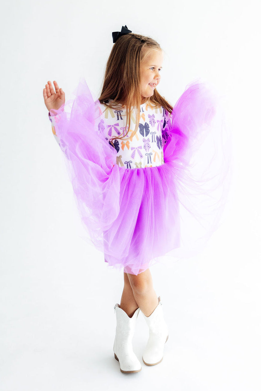A young girl wearing a whimsical purple and white costume with a patterned bodice, a voluminous purple tulle skirt, and matching purple wings, paired with white cowboy boots, set against a plain white studio background.