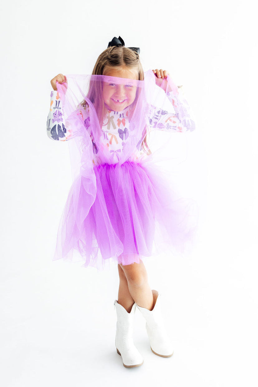 A young girl wearing a purple tulle tutu dress with a colorful patterned long-sleeve top and white cowboy boots, holding a sheer purple veil in front of her face against a white background.