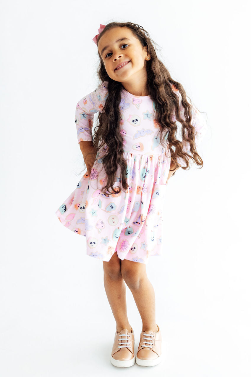 A young girl with long, wavy brown hair stands against a plain white background wearing a short-sleeved pink dress with a colorful cartoon pattern and beige sneakers.
