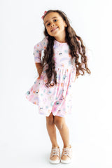 A young girl with long, wavy brown hair stands against a plain white background wearing a short-sleeved pink dress with a colorful cartoon pattern and beige sneakers.