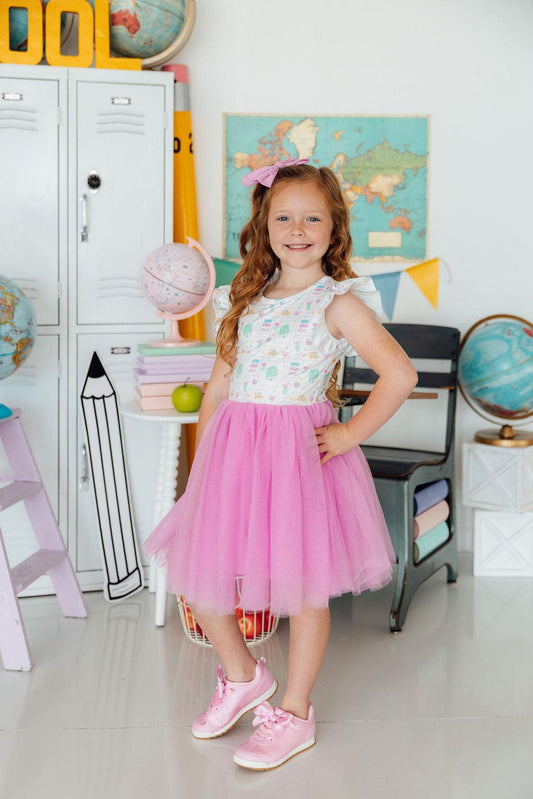 Young girl in a colorful dress with a pink skirt standing in a classroom setting.