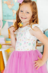 Young girl in a dress with a floral top and pink skirt, standing in a colorful room.