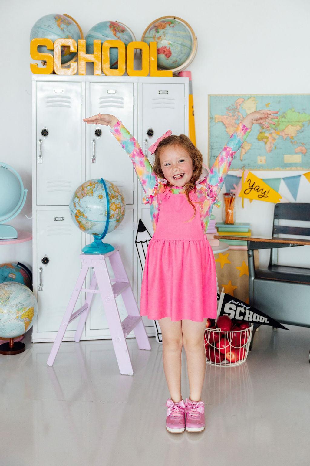 Child in a pink dress standing in a classroom with globes and lockers.