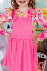 Child wearing a pink dress with colorful long-sleeve shirt underneath, standing indoors.