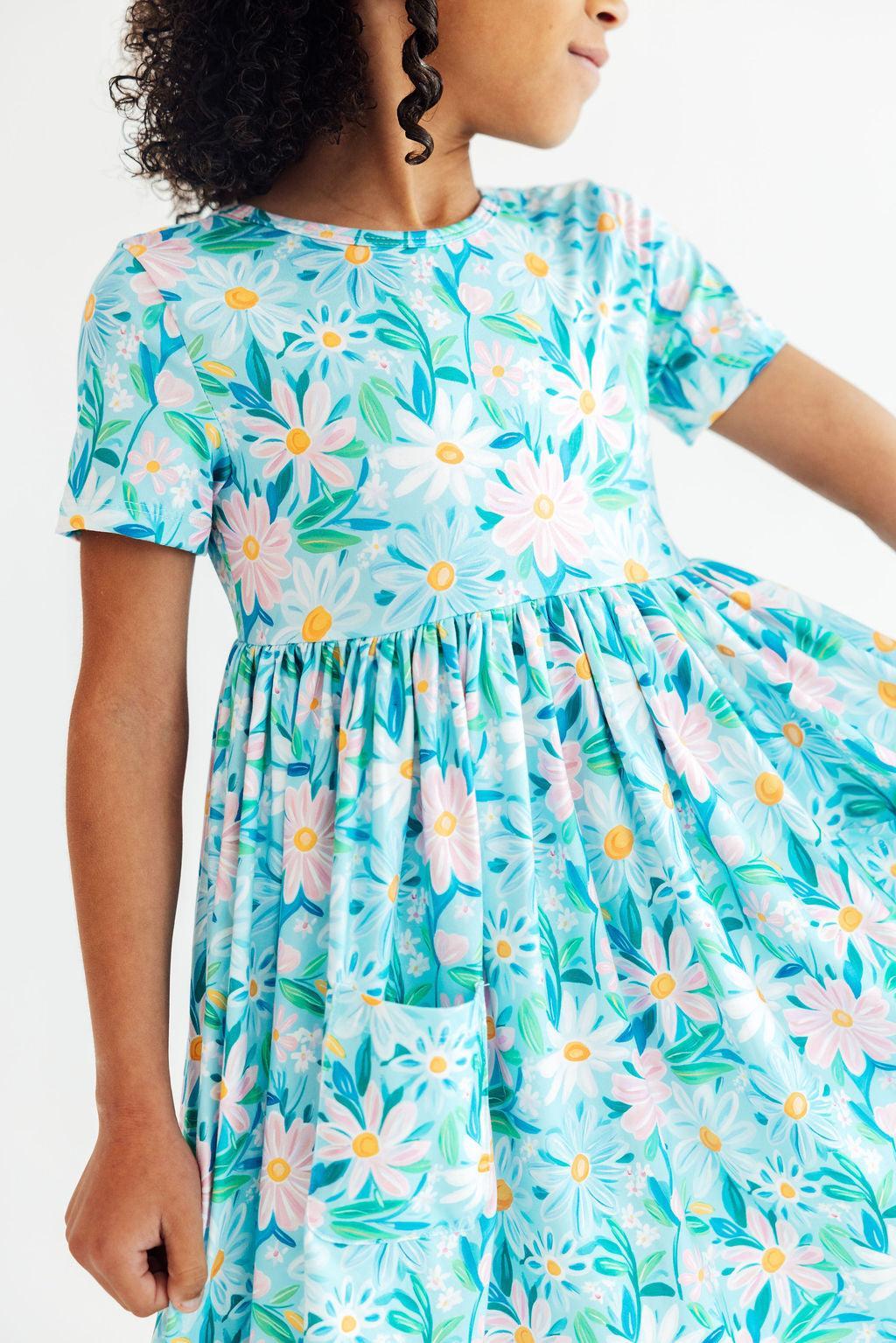 A child wearing a short-sleeve dress with a light blue background and a colorful floral print featuring daisies and other flowers in pink, yellow, green, and white. The dress has a gathered waist and a front patch pocket.