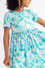 A child wearing a short-sleeve dress with a light blue background and a colorful floral print featuring daisies and other flowers in pink, yellow, green, and white. The dress has a gathered waist and a front patch pocket.